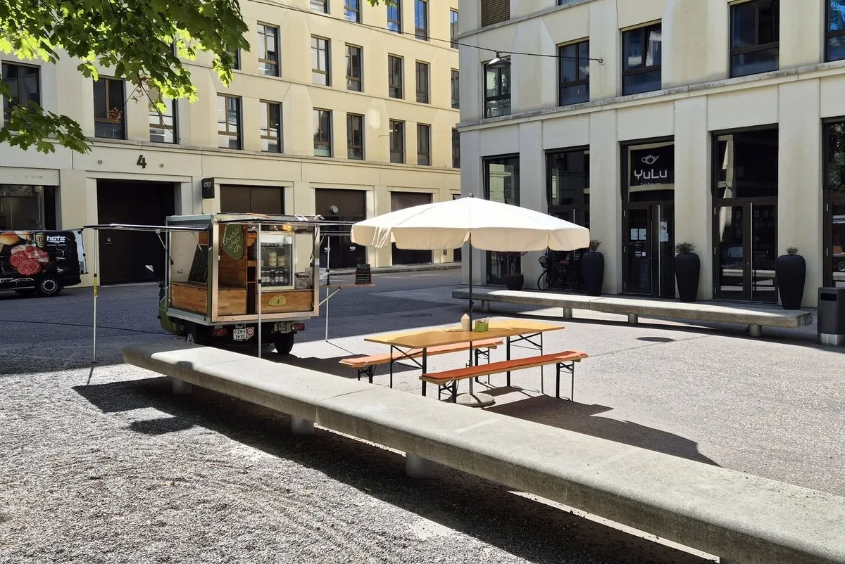 Beliebter Treffpunkt: Reto Günthards Foodtruck brachte jeweils über Mittag etwas Leben in das Neubaugebiet beim Bahnhof Uster. Der Piaggo Ape von Chabis-Chäs steht vor modernen Häusern. Davor ein weisser Sonnenschirm und eine Festbank.